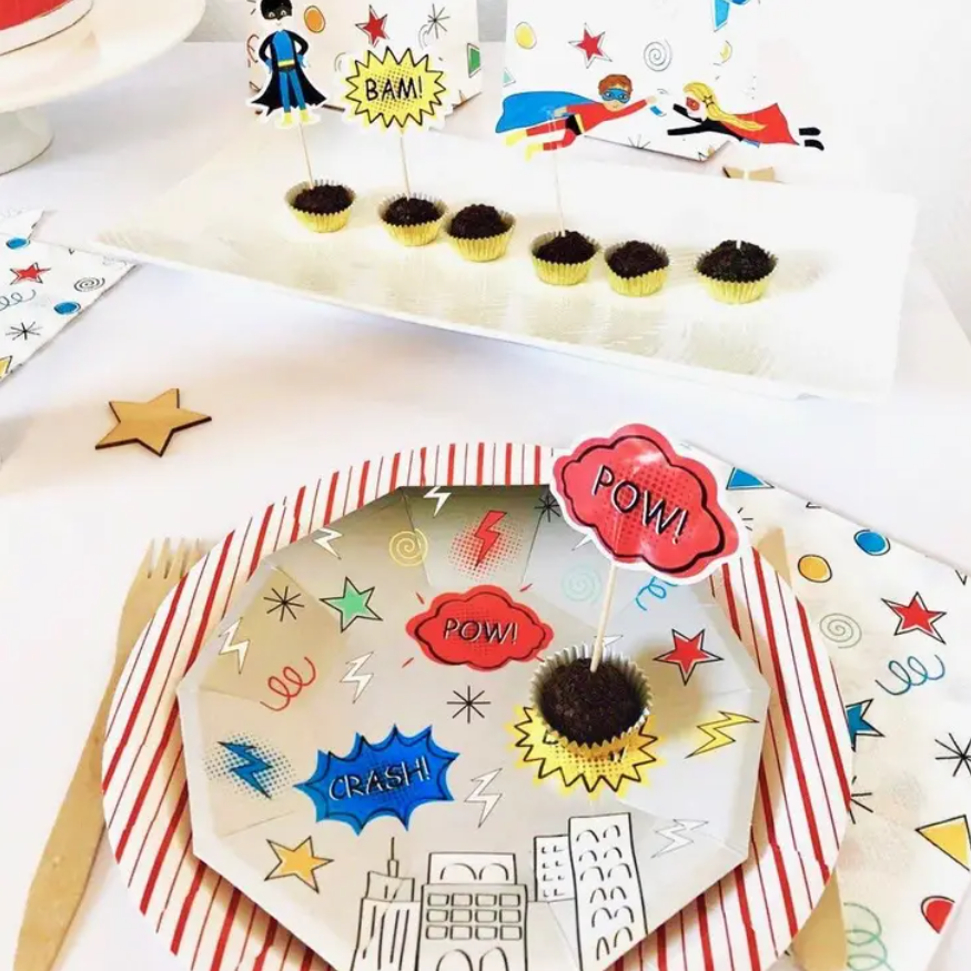 Pratos e guardanapos com padrão de banda desenhada colorida e cupcakes decorados com onomatopeias.