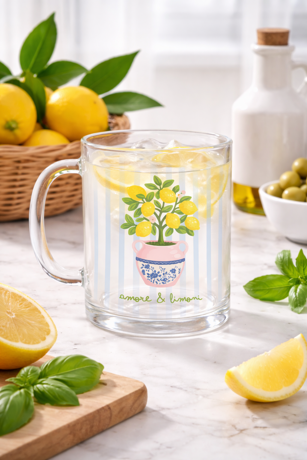 Caneca de vidro transparente com ilustração de limoeiro e texto amore & limoni contendo bebida com fatias de limão e gelo numa mesa com limões, manjericão, azeitonas e garrafa.