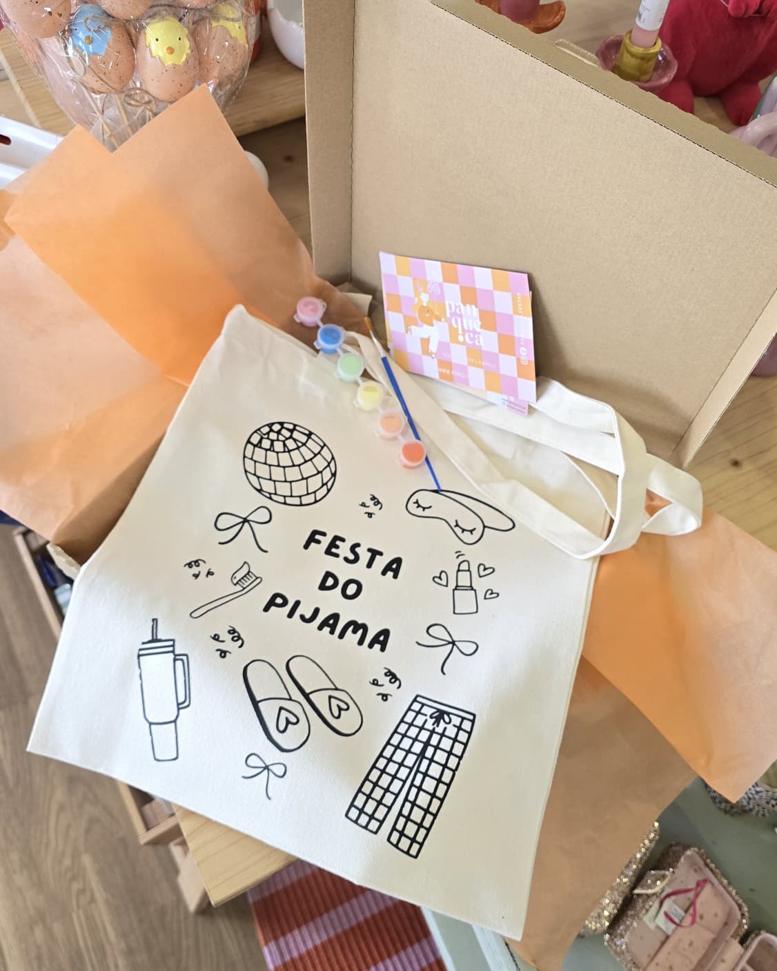 Saco de pano branco com ilustrações e texto FESTA DO PIJAMA e materiais de pintura sobre uma mesa.