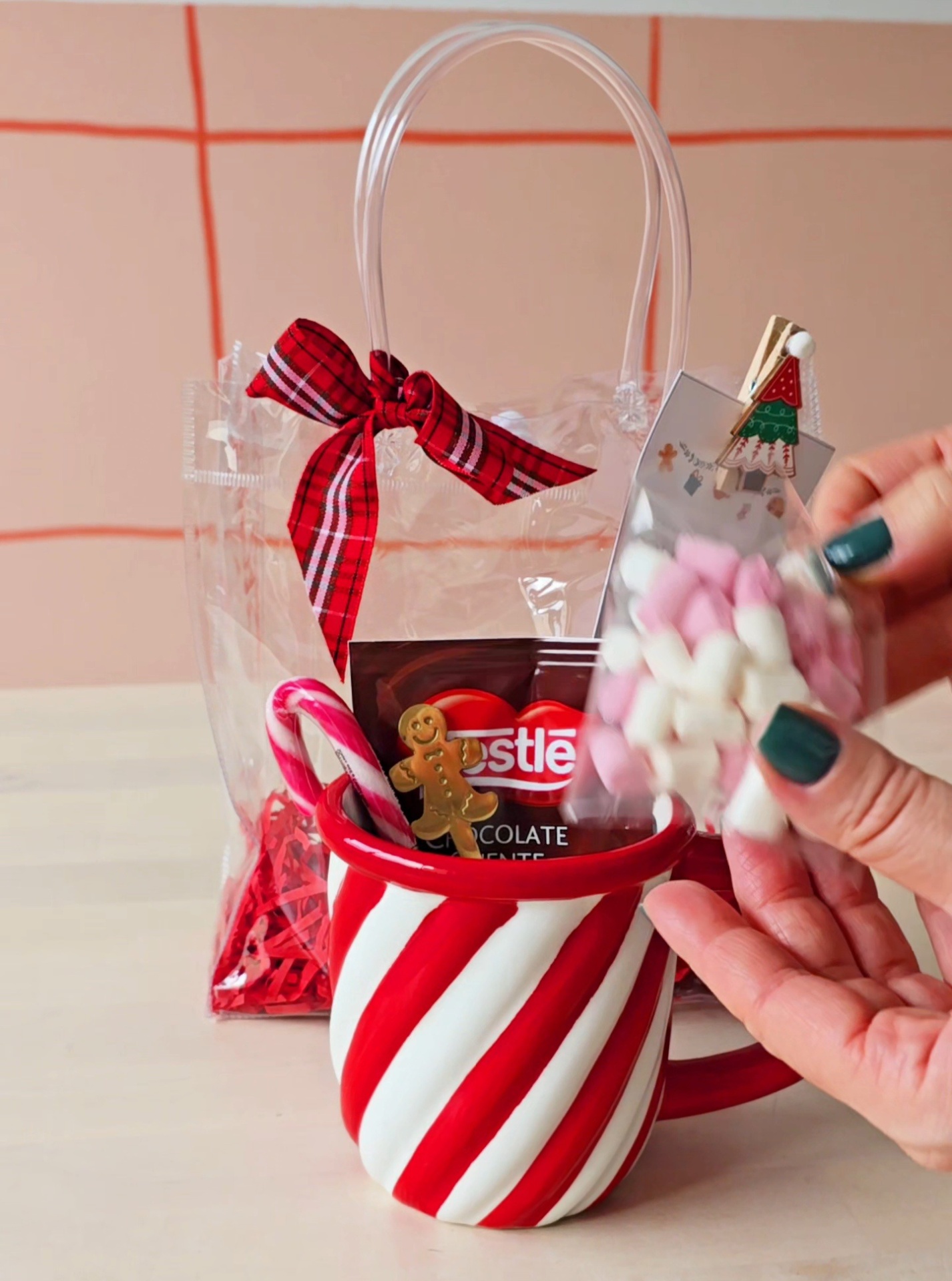 Caneca vermelha e branca com chocolate Nestlé e enfeites dentro, saco transparente com laço vermelho atrás.