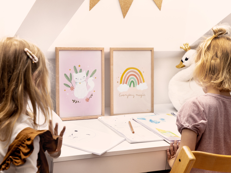 Dois quadros infantis com moldura de madeira em mesa branca, crianças desenhando ao redor