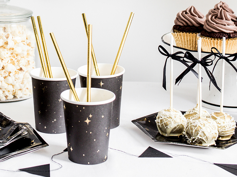 Copos de papel pretos com estrelas douradas e canudos dourados, cupcakes com cobertura castanha, bolos brancos em palitos e recipiente com pipocas numa mesa decorada a preto e branco.