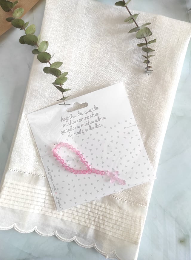 Toalha creme com bainha ondulada e cartão com pulseira rosa num saco plástico sobre a toalha