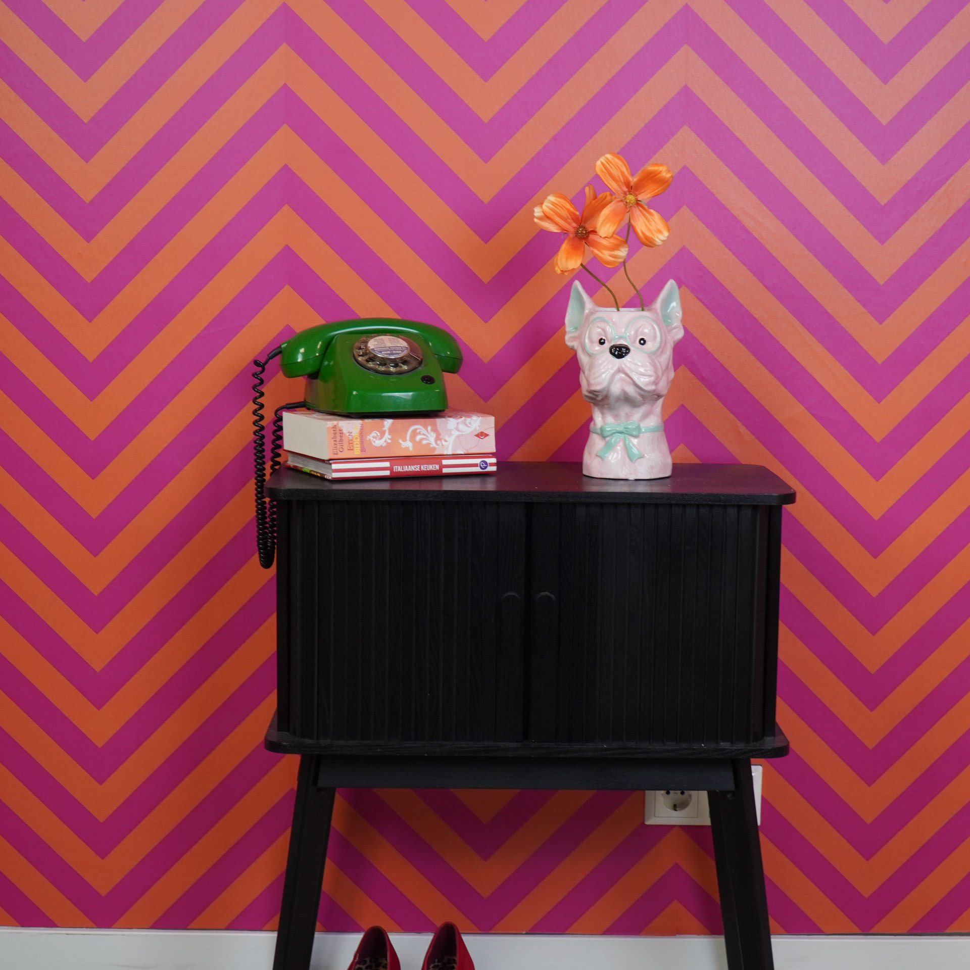 Móvel preto com telefone verde, vaso cão rosa com flores e livros, fundo de parede zig zag laranja e rosa.