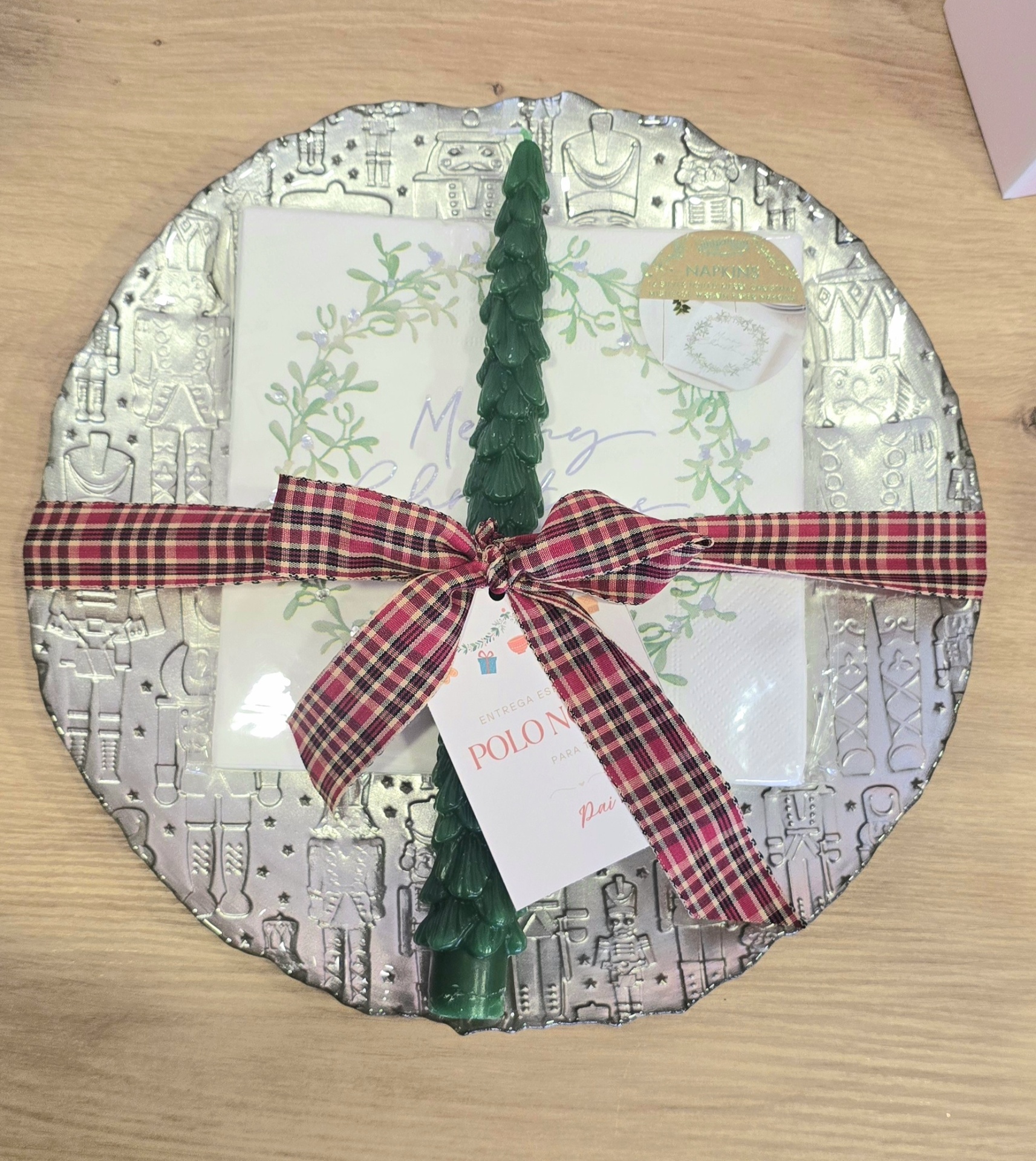 Conjunto de guardanapos de Natal com motivos decorativos, prato prateado com desenhos, laço xadrez e vela verde em forma de árvore de Natal.