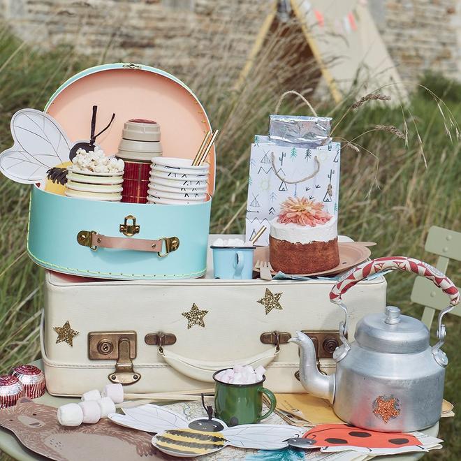 Mesa com malas vintage, bolo decorado, chaleira metálica e acessórios de festa ao ar livre