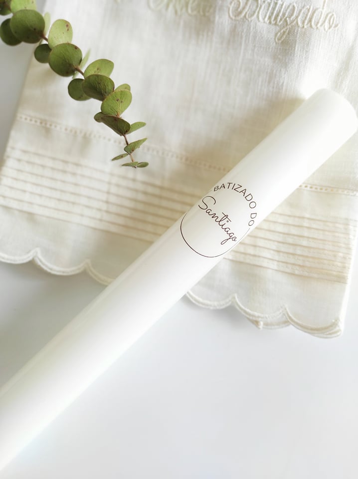 Vela branca com etiqueta personalizada sobre tecido branco bordado e folhas verdes