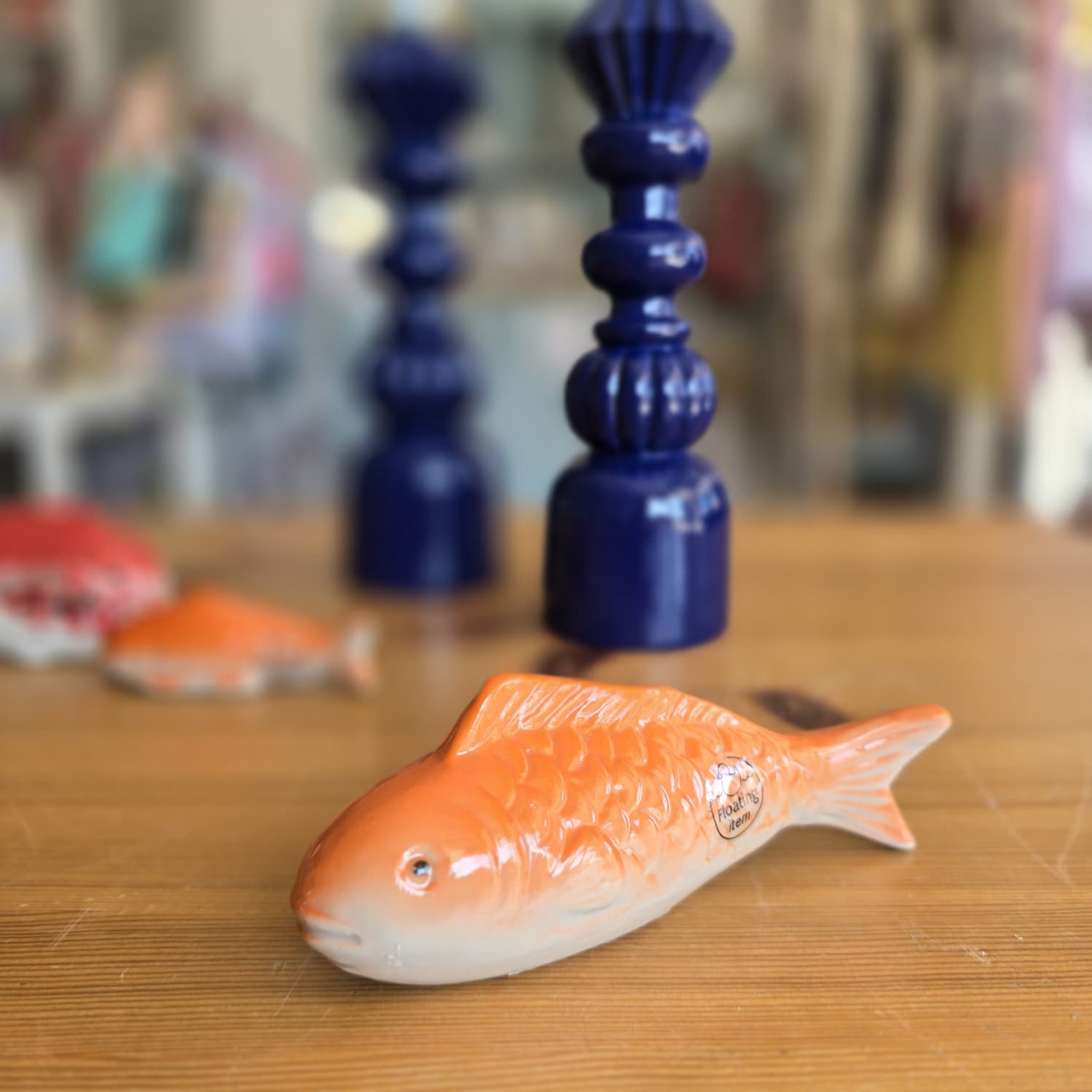 Peixe decorativo de cerâmica laranja sobre mesa de madeira com castiçais azuis no fundo