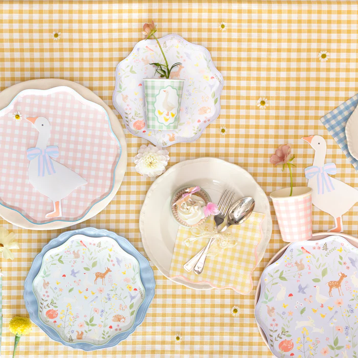 Mesa posta com toalha xadrez amarela e pratos e copos com desenhos de gansos e flores.