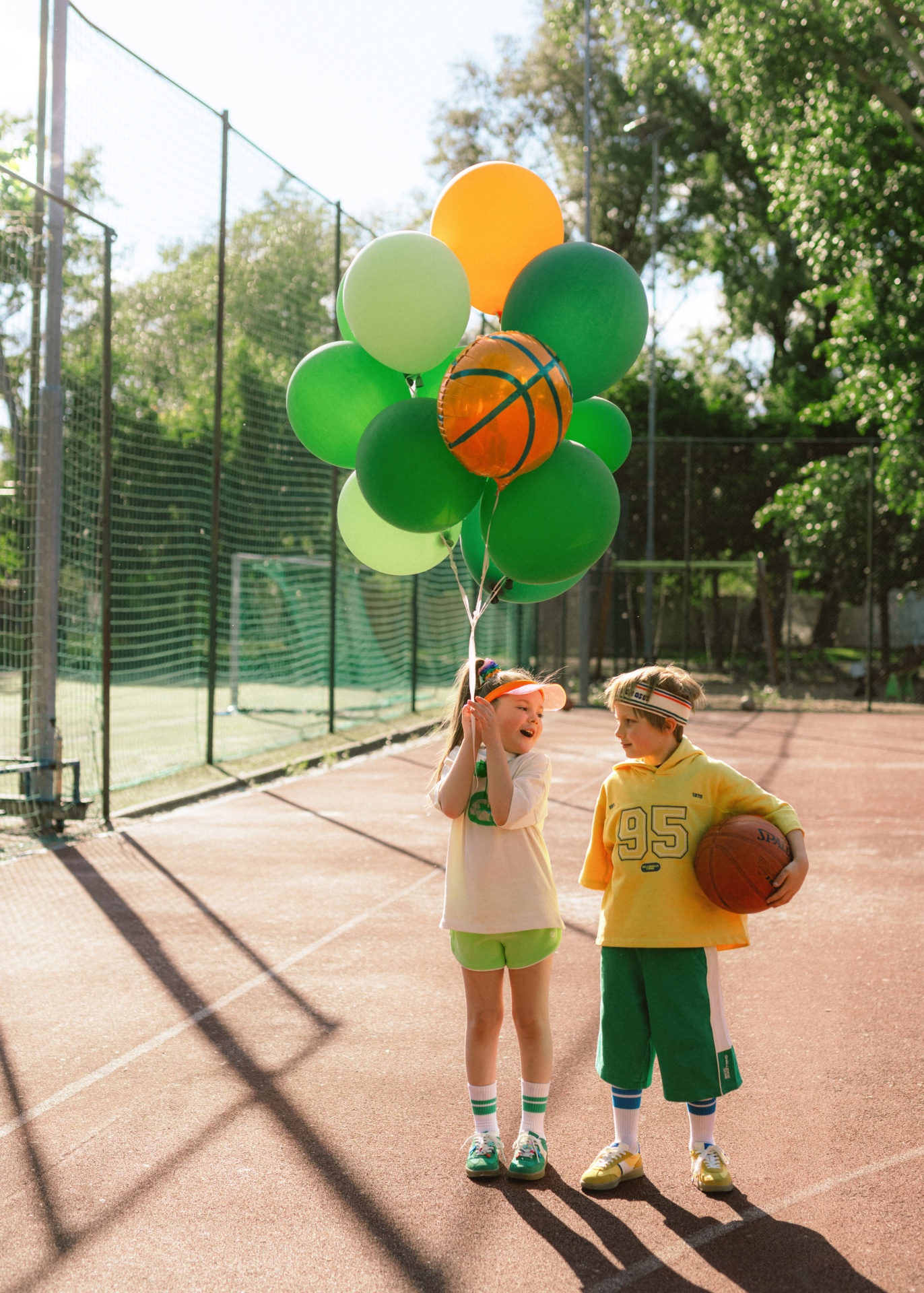 Duas crianças numa quadra desportiva com balões coloridos e bola de basquetebol