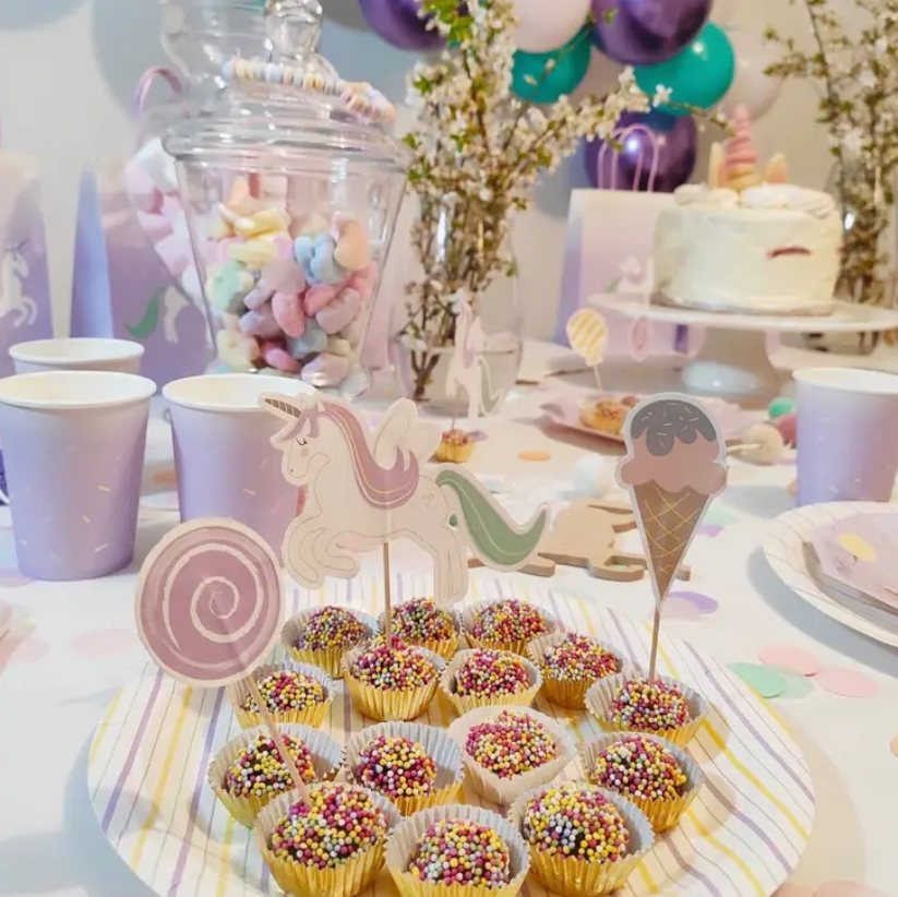 Mesa festiva com brigadeiros coloridos, copos lilás, bolo branco e decoração com marshmallows e balões