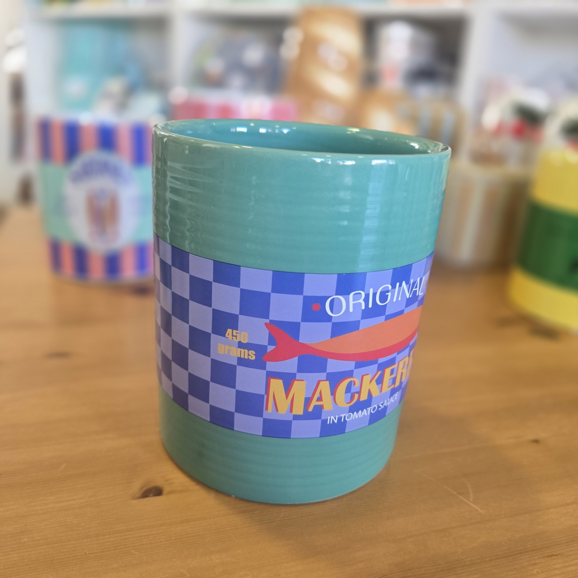 Caneca azul esverdeada com etiqueta azul e branca com texto e peixe, sobre mesa de madeira.