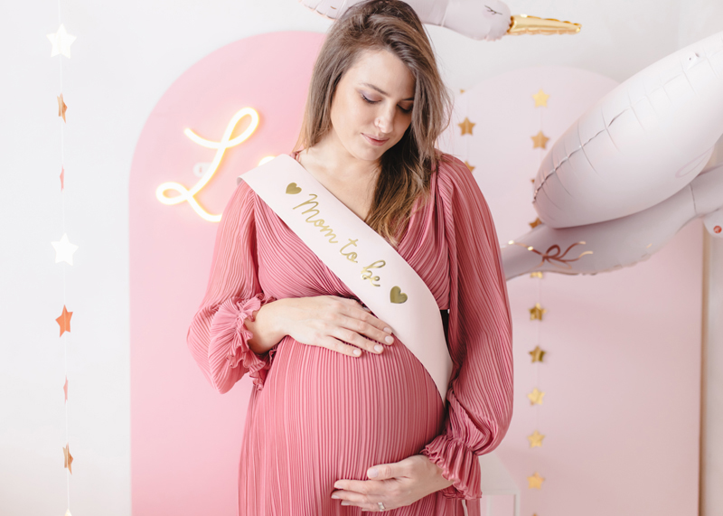 Mulher grávida com vestido rosa e faixa 'Mom to Be' em ambiente decorado com balões e estrelas.