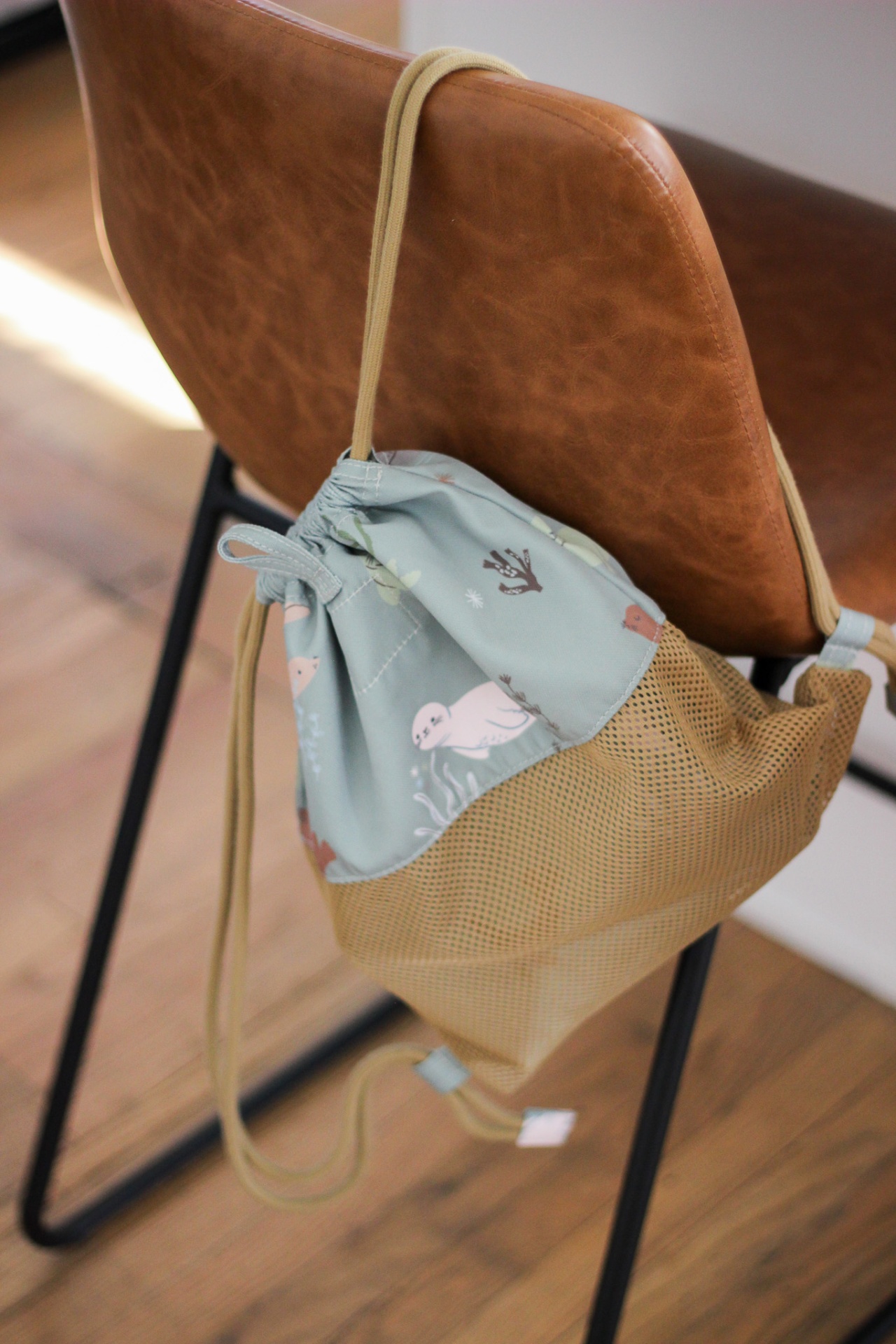 Mochila com cordão azul-claro estampada com animais marinhos e parte inferior em malha bege pendurada numa cadeira de pele castanha.