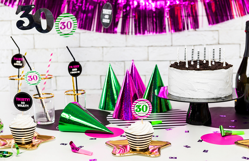 Decoração de festa com chapéus de cone verdes e roxos, bolo branco com velas, copos com canudos decorativos 30, mesa branca e fundo com cortina roxa metálica.