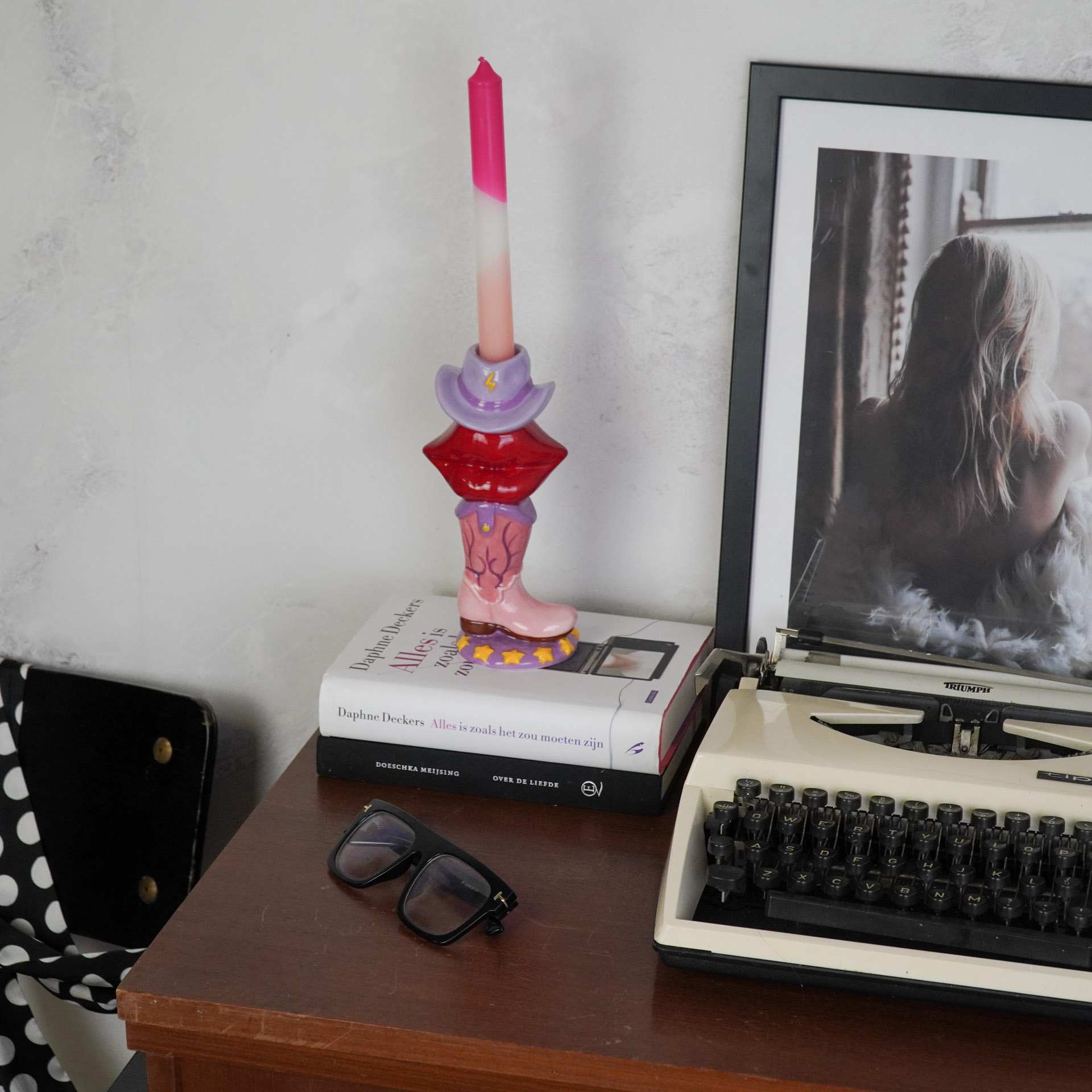 Castiçal colorido em forma de bota com vela rosa sobre livros numa mesa com óculos e máquina de escrever.
