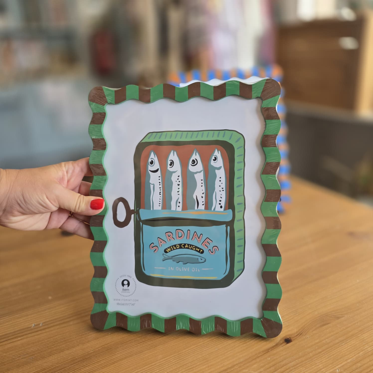 Quadro decorativo com moldura ondulada verde e castanha, ilustração de lata de sardinhas e texto em inglês.