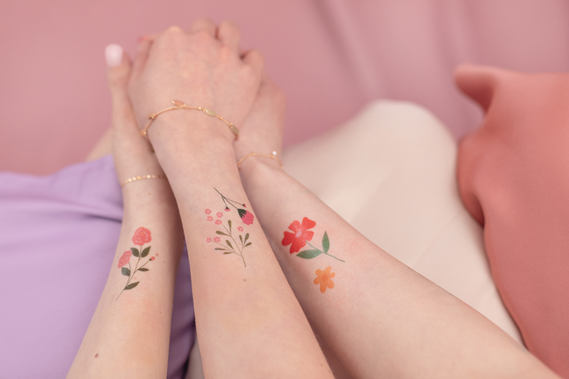 Braços com tatuagens temporárias florais coloridas e pulseiras delicadas sobre tecidos coloridos.