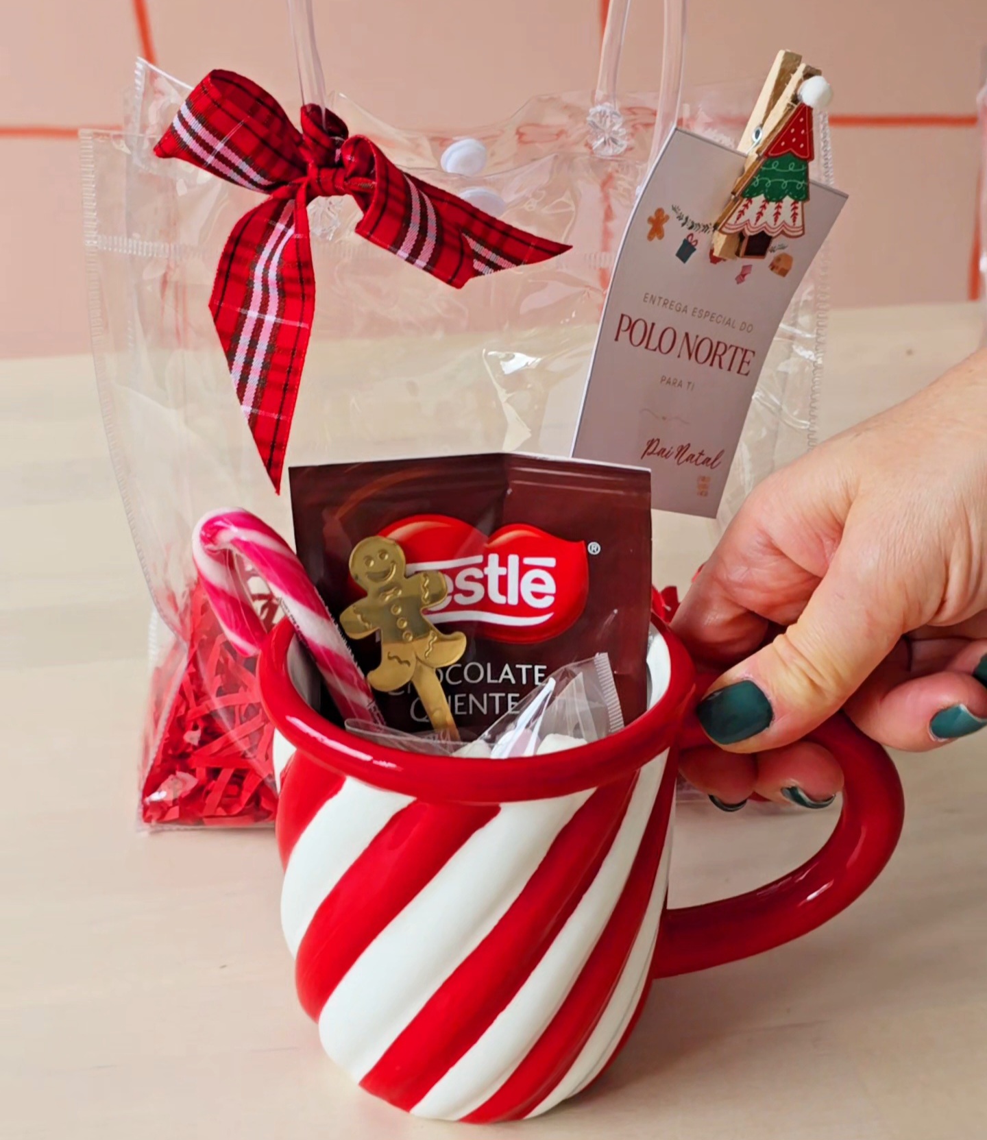 Caneca listrada vermelha e branca com chocolate Nestlé e acessórios em fundo neutro com saco transparente e laço