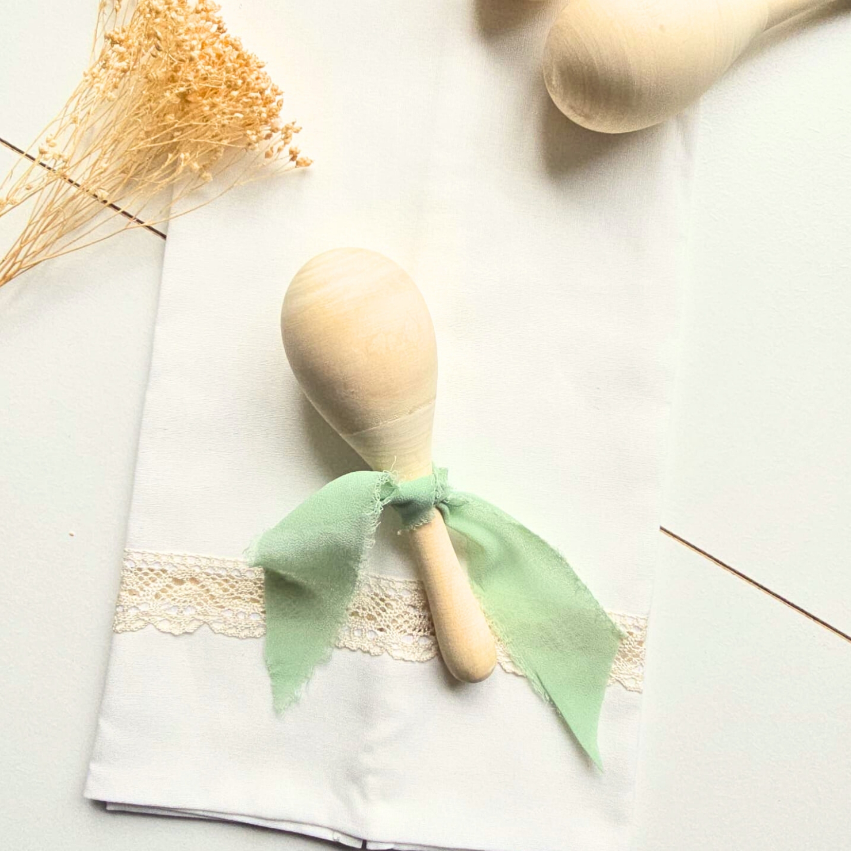 Maraca de madeira clara com laço verde sobre pano branco com renda, flores secas ao lado