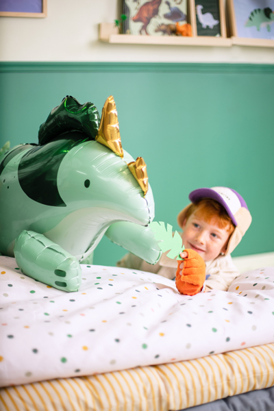 Balão de dinossauro verde e dourado com criança segurando brinquedo em cama colorida