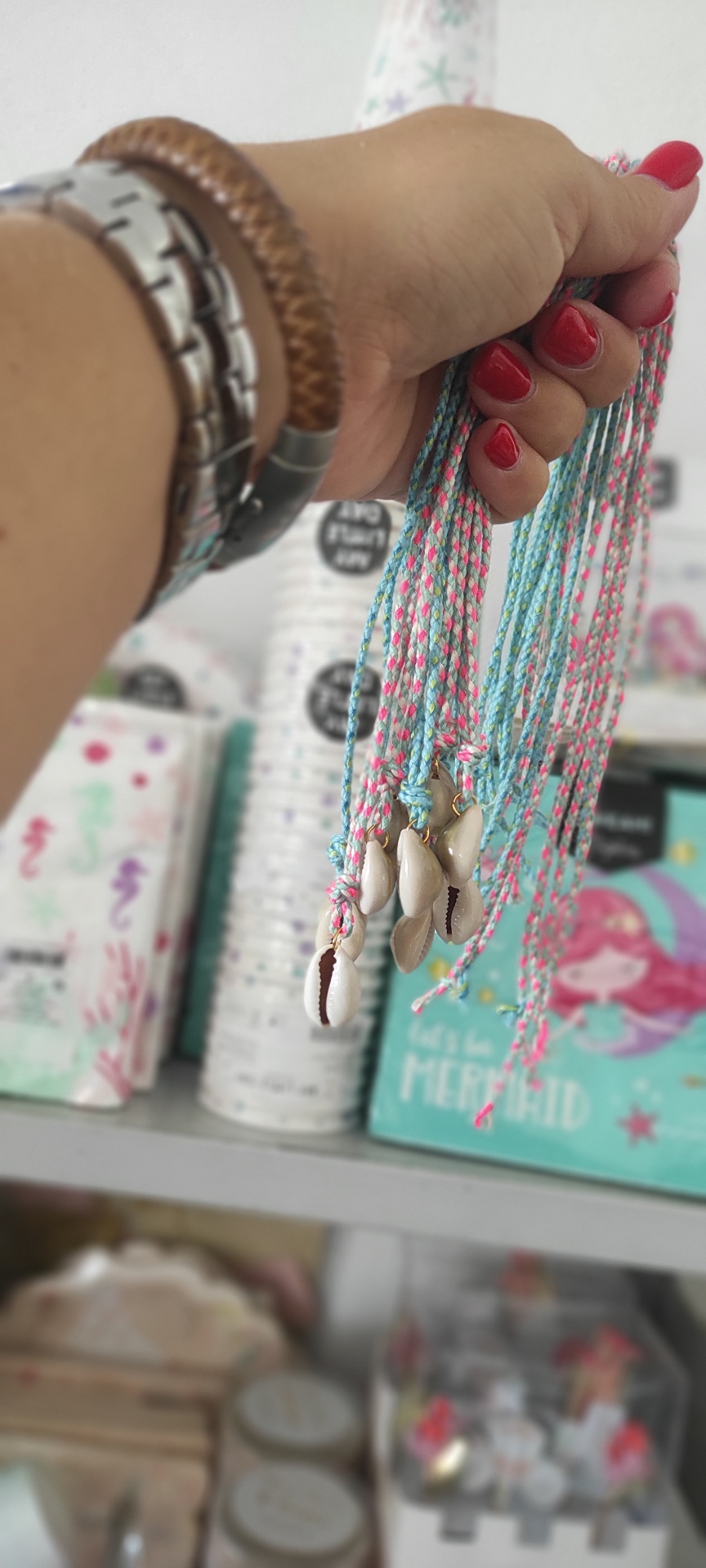 Colares com conchas em cordões coloridos segurados por mão feminina com fundo de caixas coloridas com tema de sereia