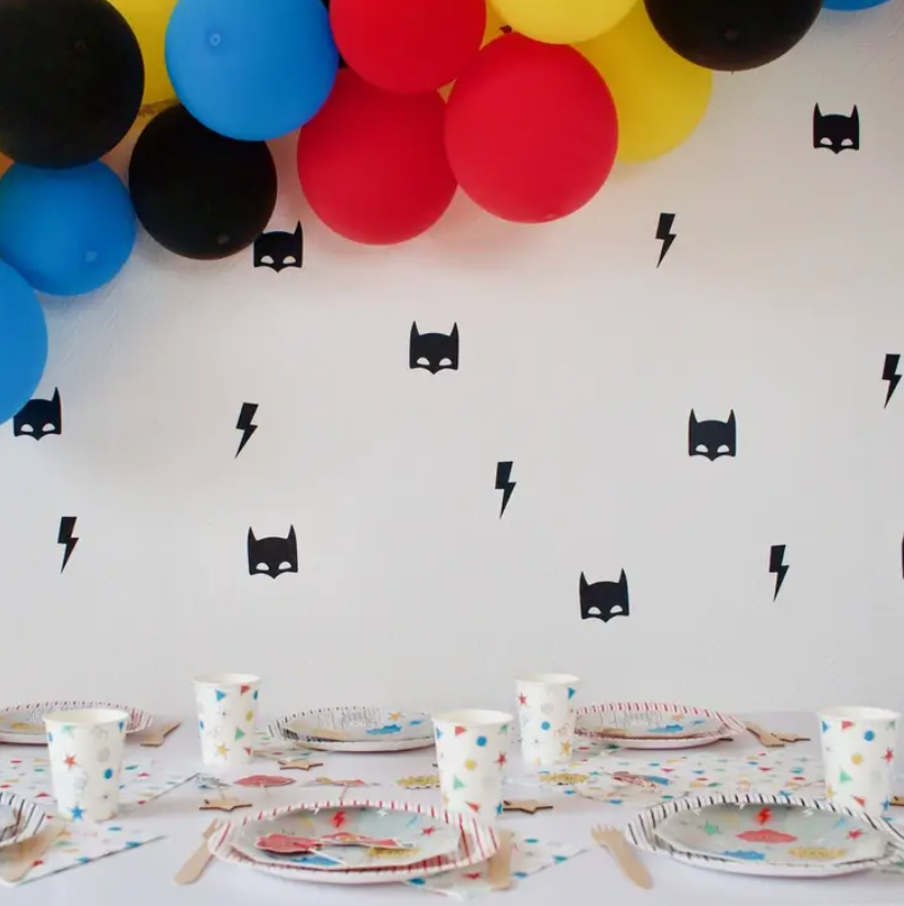 Decoração de festa com balões coloridos, pratos e copos descartáveis com padrão, talheres de madeira e autocolantes de Batman na parede