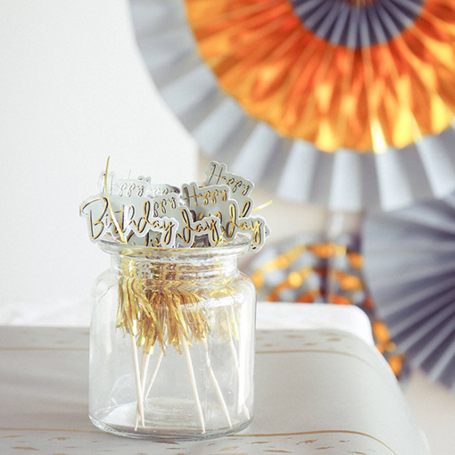 Frasco de vidro com toppers dourados de 'Happy Birthday' e decorações coloridas de papel ao fundo