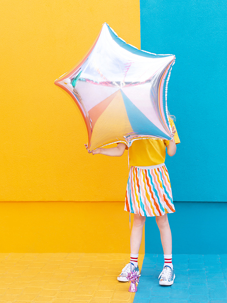 Pessoa segurando balão estrela prateado com roupa colorida em fundo amarelo e azul