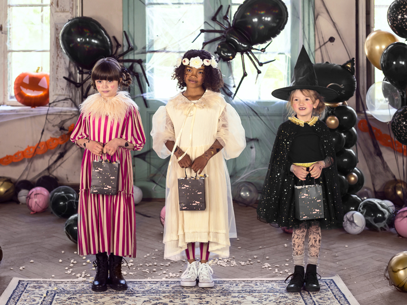 Três crianças em fatos de Halloween segurando sacos pretos, decoradas com balões e abóbora de Halloween ao fundo.