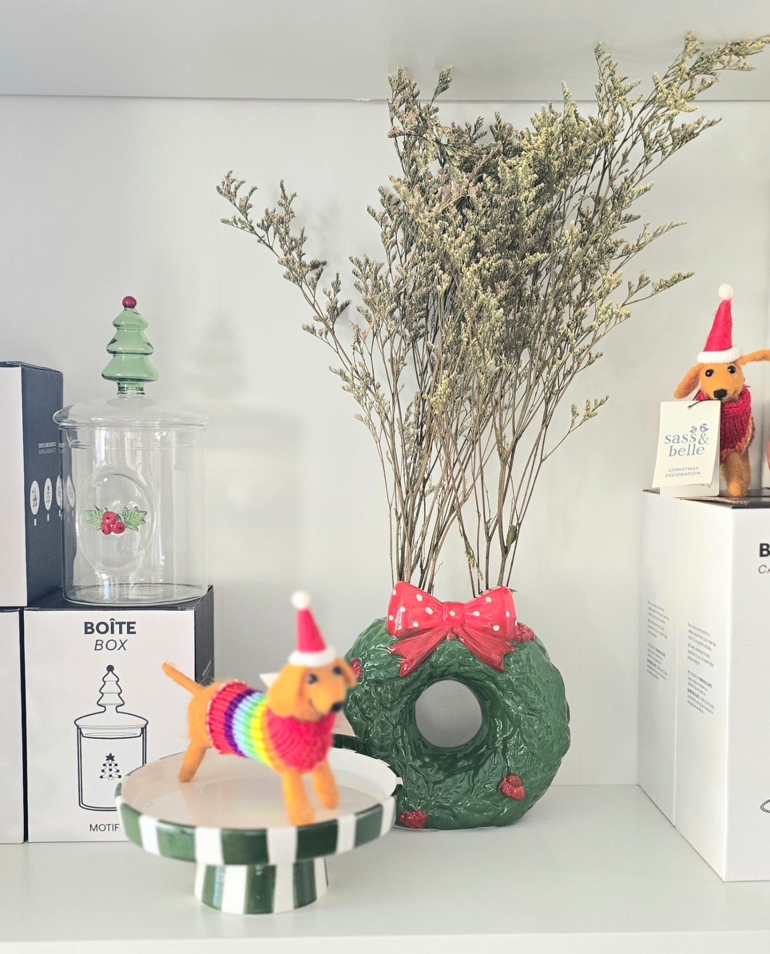 Decoração de Natal com vaso em forma de coroa, boneco de cão com gorro de Natal e caixa na prateleira.