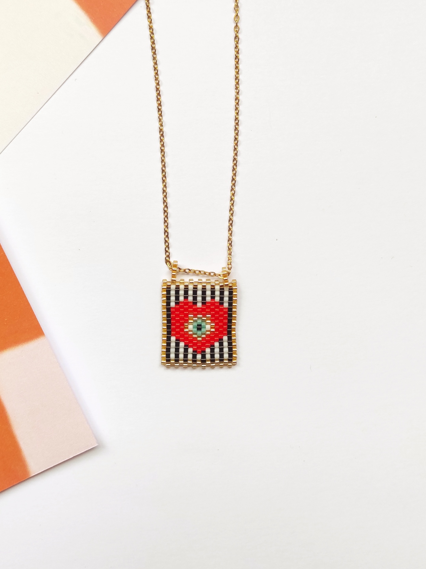 Colar dourado com pendente retangular de contas coloridas com coração vermelho.