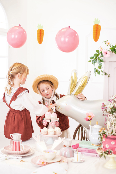 Duas meninas com roupas cor de ferrugem e branco junto a mesa decorada com chávenas e balões rosa e de coelho