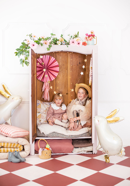 Armário rosa vintage decorado com flores e luzes, duas crianças sentadas no seu interior, coelhos decorativos ao lado.