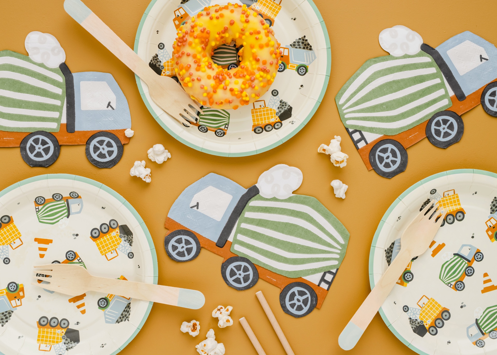 Mesa com pratos descartáveis de papel e guardanapos de camião de cimento, donuts com cobertura amarela, forquilha de madeira e pipocas.