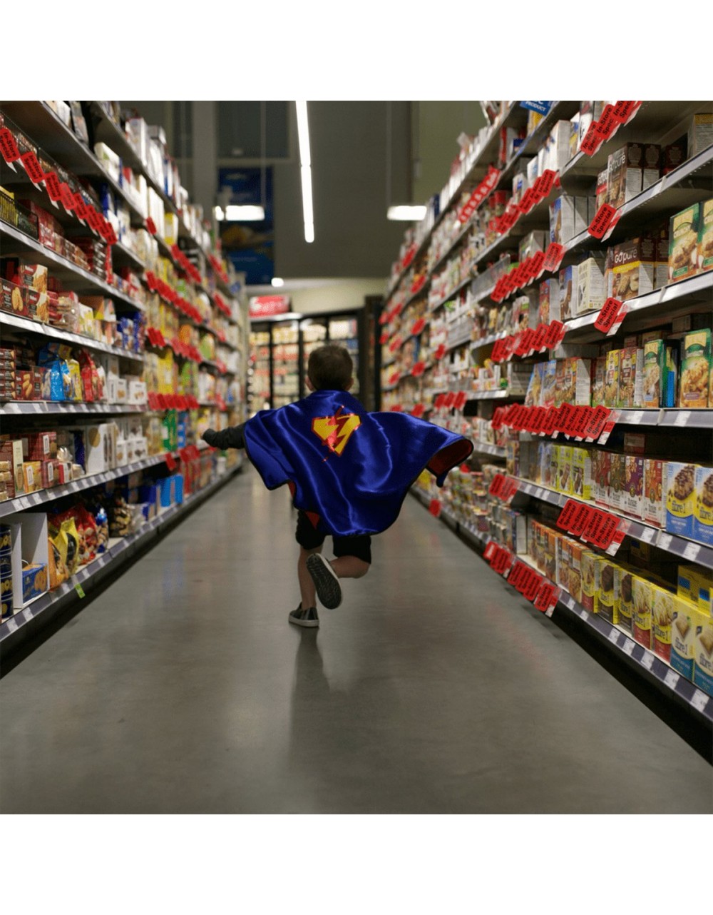 Corredor de supermercado com prateleiras cheias de produtos embalados e criança com capa azul correndo no centro.