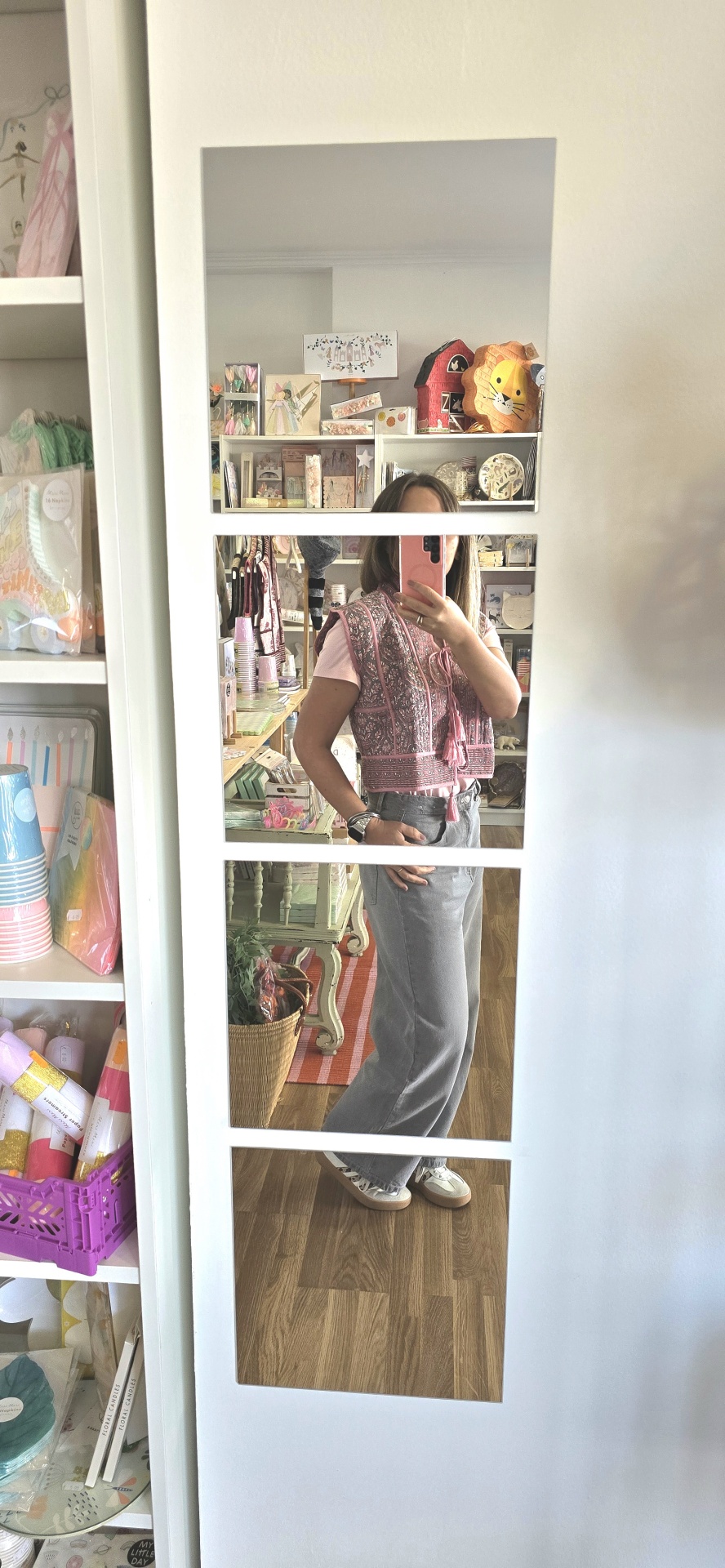 Pessoa vestindo roupa rosa e cinza refletida em espelho branco dividido em quatro partes com prateleiras e decoração ao fundo