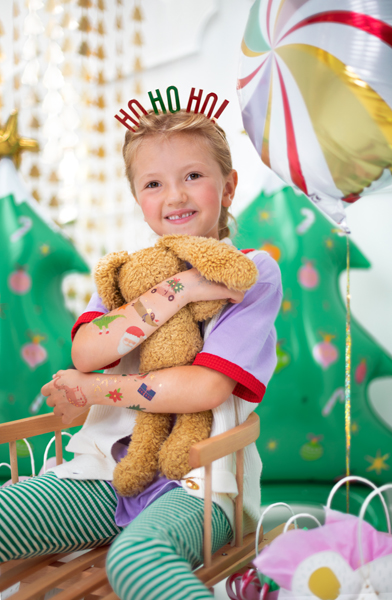 Criança com diadema 'HO HO HO' abraçando urso de peluche com decoração natalícia ao fundo