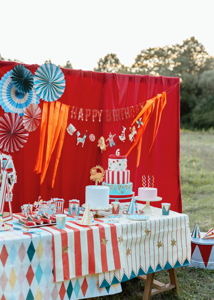 Mesa de aniversário decorada com bolo temático de circo, toalhas coloridas e chapéus de festa num espaço exterior