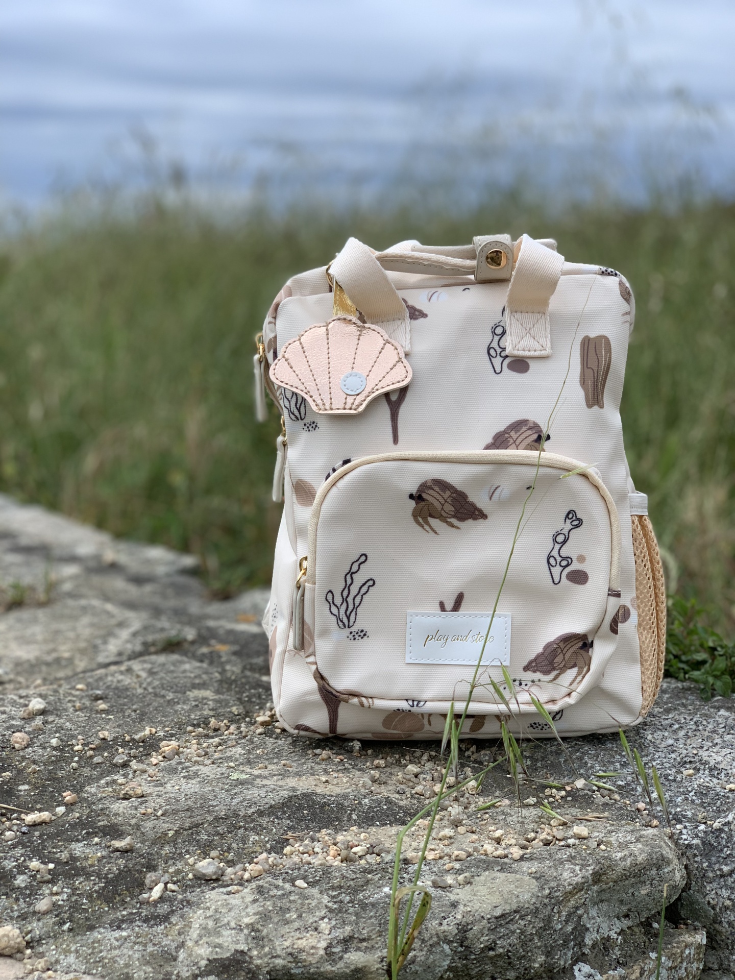 Mochila pequena bege com padrão de algas e conchas, bolso frontal, etiqueta PLAYGROUND e porta-chaves em forma de concha rosa, sobre pedra exterior