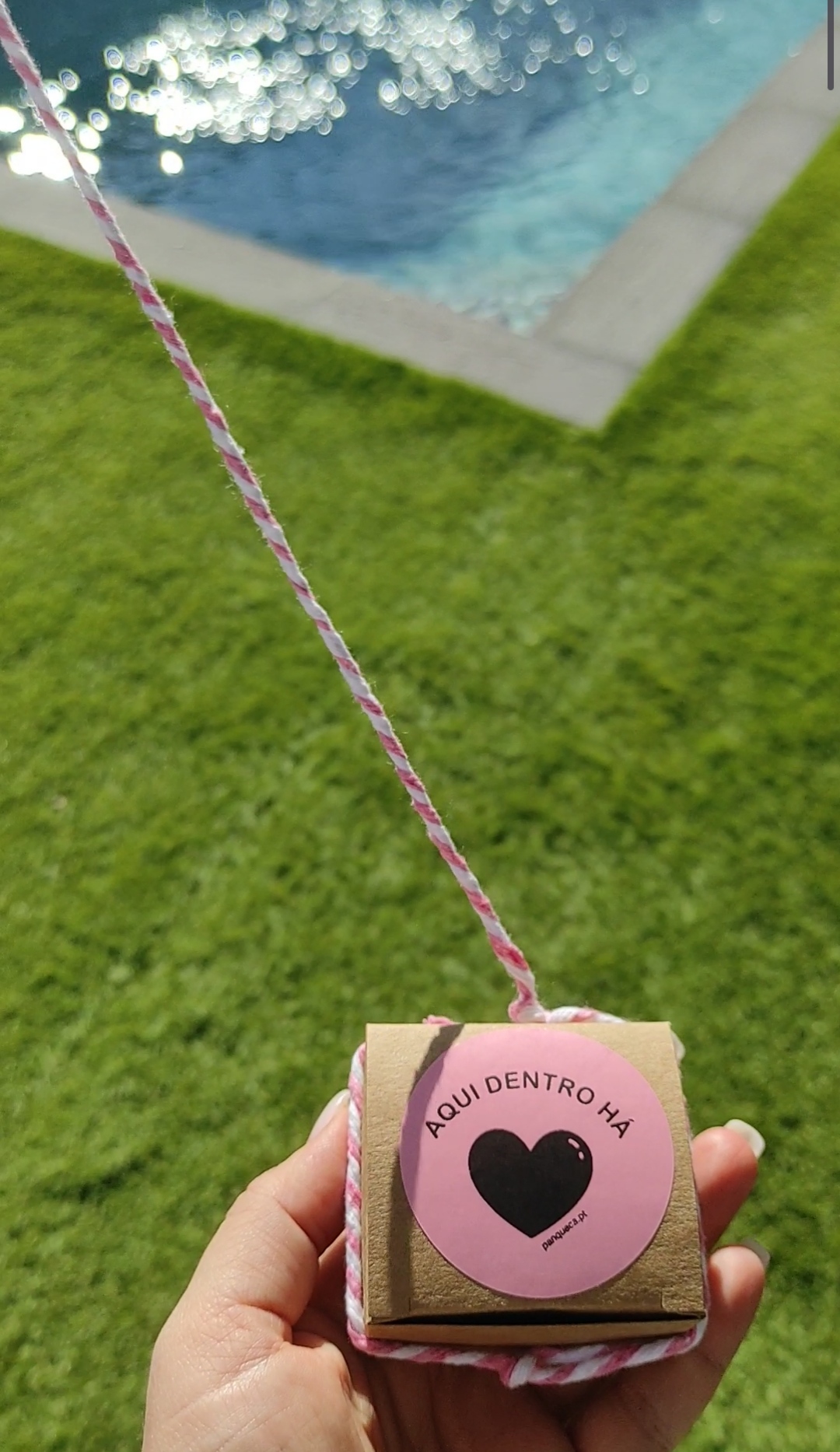 Caixa pequena de cartão com etiqueta rosa e cordel colorido perto de piscina e relvado