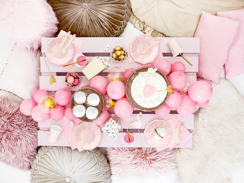 Mesa baixa cor-de-rosa decorada com balões e acessórios cor-de-rosa, rodeada por almofadas e tapetes pastel