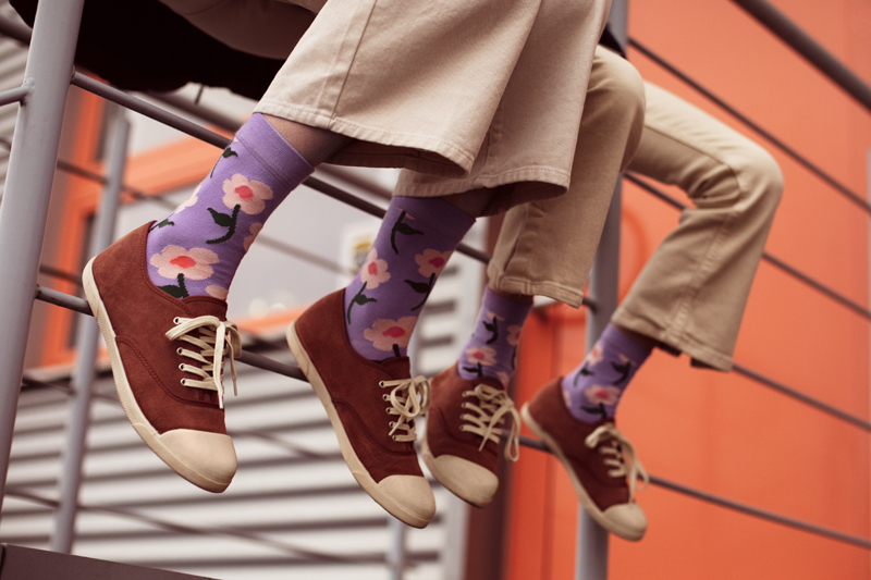 Pessoas com meias roxas com flores coloridas e sapatos castanhos sentadas numa estrutura metálica junto a parede laranja.