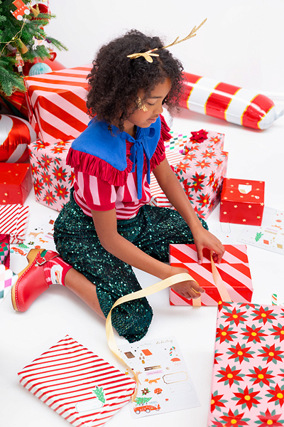 Menina a embrulhar presente com papel riscado vermelho e branco, rodeada de presentes coloridos