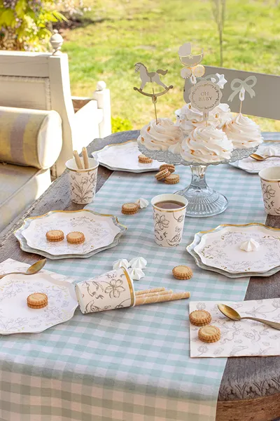 Mesa posta com toalha xadrez azul e branca, pratos e copos florais, bolachas e cupcakes decorados com toppers OH BABY.