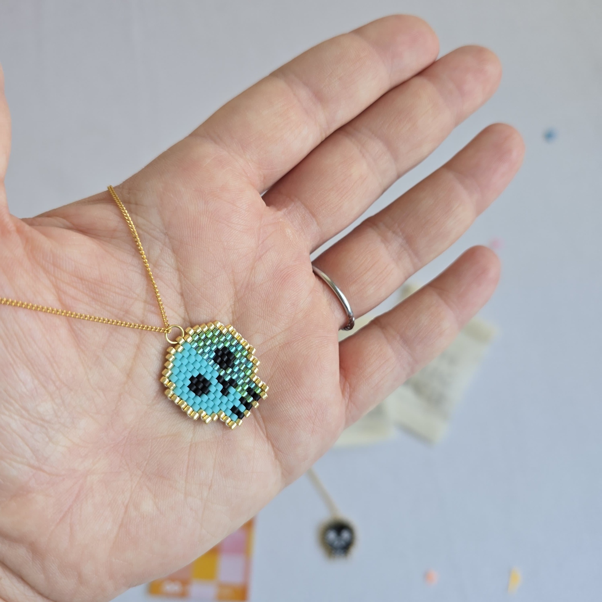 Colar com pendente de contas formando uma caveira azul e preta segurado por mão com anel.