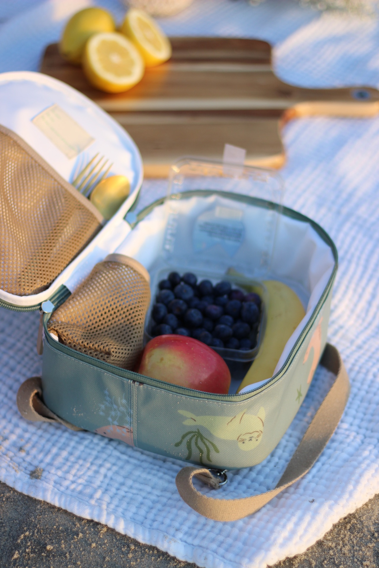 Saco térmico verde água com frutas e interior branco aberto sobre toalha branca ao ar livre