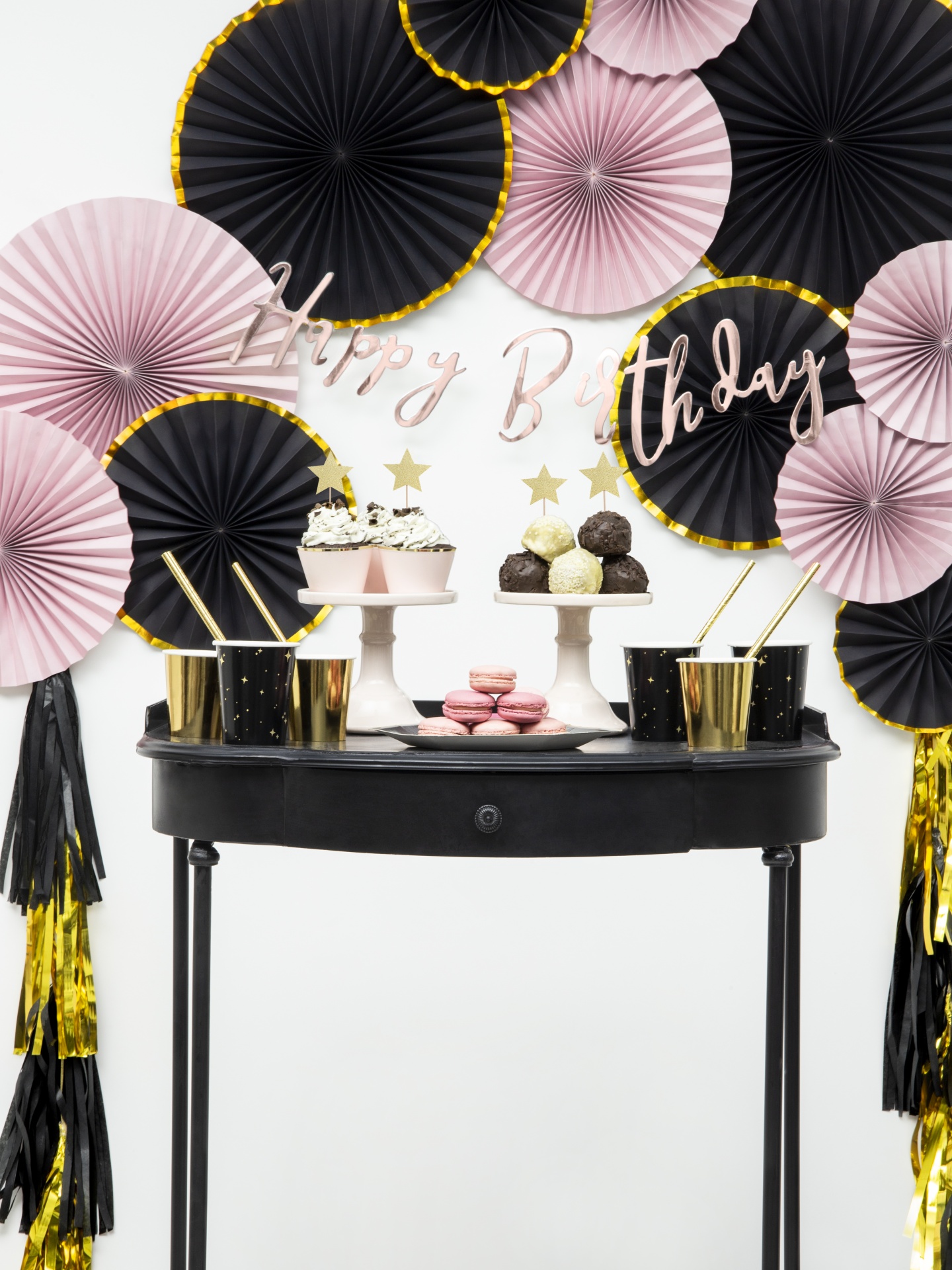 Decoração de festa de aniversário com doces e painéis de papel em rosa, preto e dourado sobre mesa preta