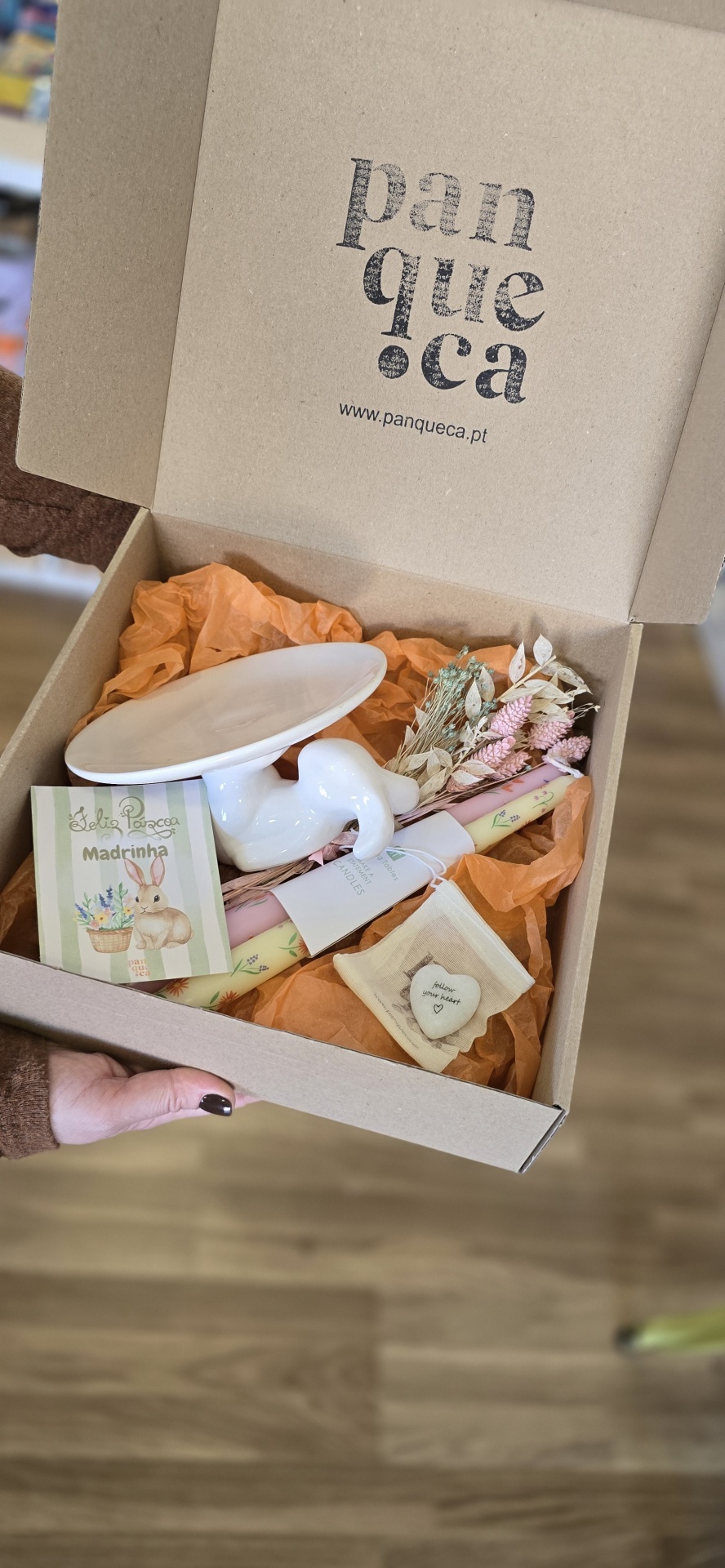 Caixa de presente com prato branco em forma de pássaro, flores secas, velas coloridas, pedra com texto em saco de tecido e cartão de felicitações com coelho