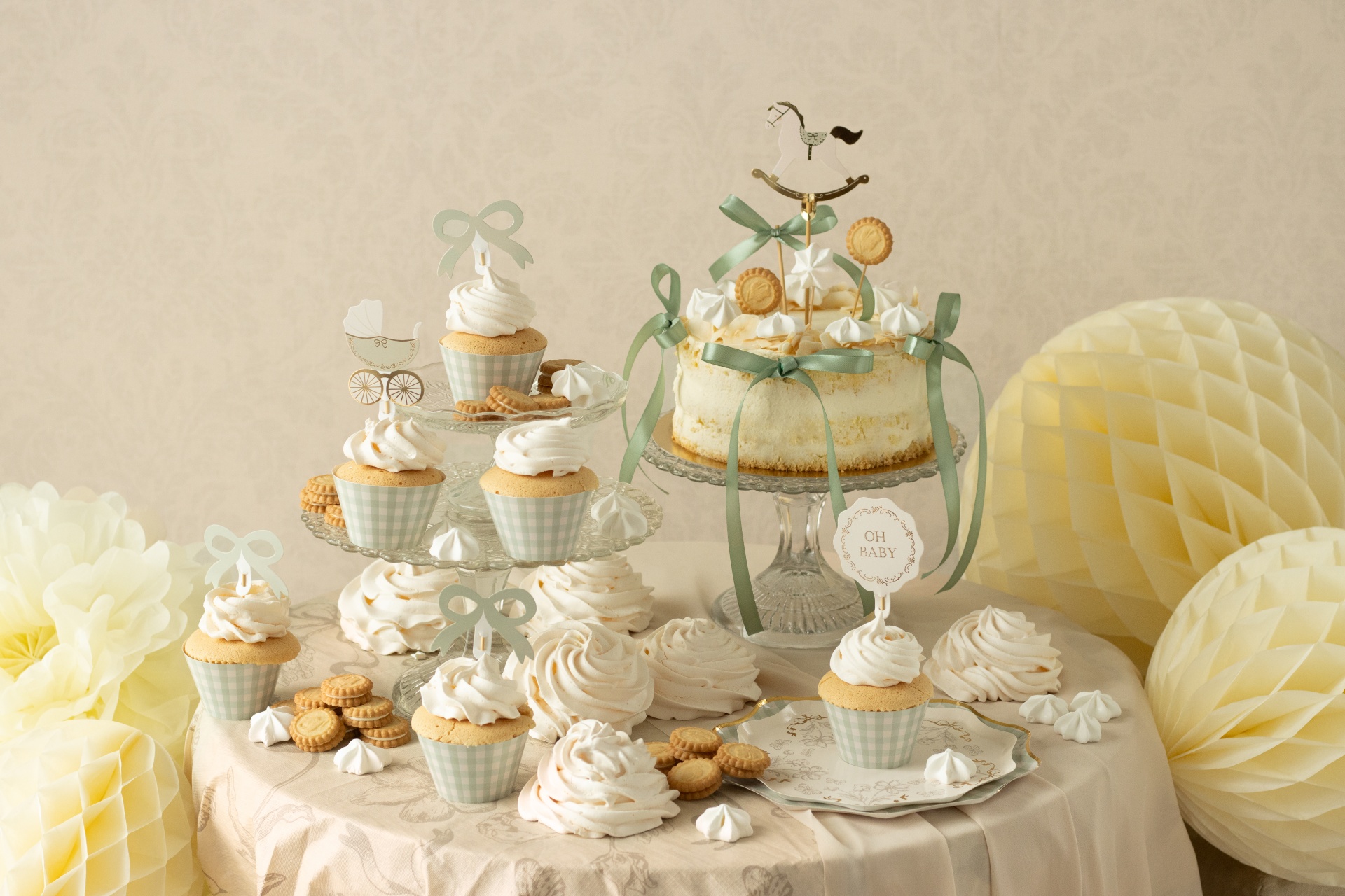 Bolo decorado com laços verdes, cupcakes com chantilly e toppers, meringues e bolachas numa mesa com toalha creme